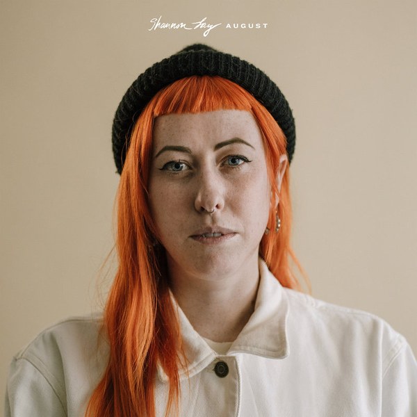 Album artwork for Shannon Lay's album, artwork. Female presenting person in a cream button up shirt, with bright orange hair and black hat looking directly at the camera with a tan background.